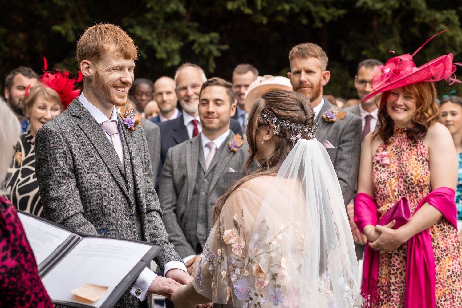 Outdoor wedding ceremony with groom in gray suit exchanging vows with bride in lace veil, guests smiling in background.