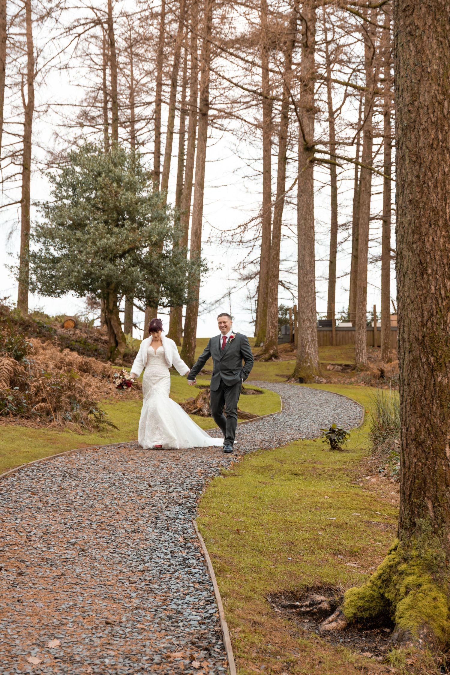 Bride and groom walk hand in hand along a winding gravel path through tall pine trees on their wedding day.