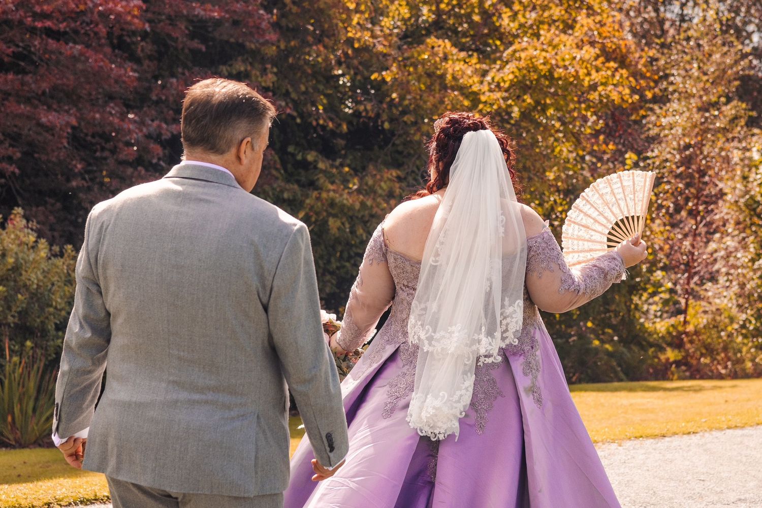 Bride in purple gown with veil and fan walks with groom in gray suit through colorful autumn garden.
