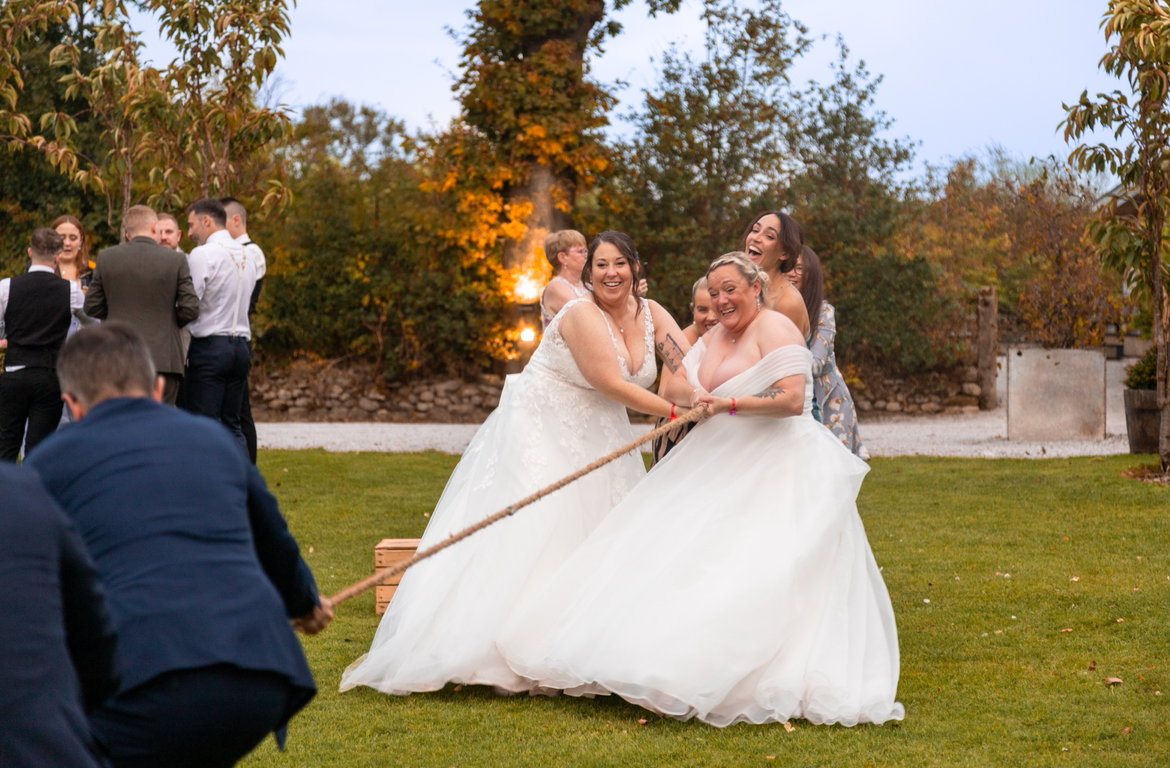 Two brides in white gowns laugh while playing tug-of-war with guests at an outdoor wedding reception.