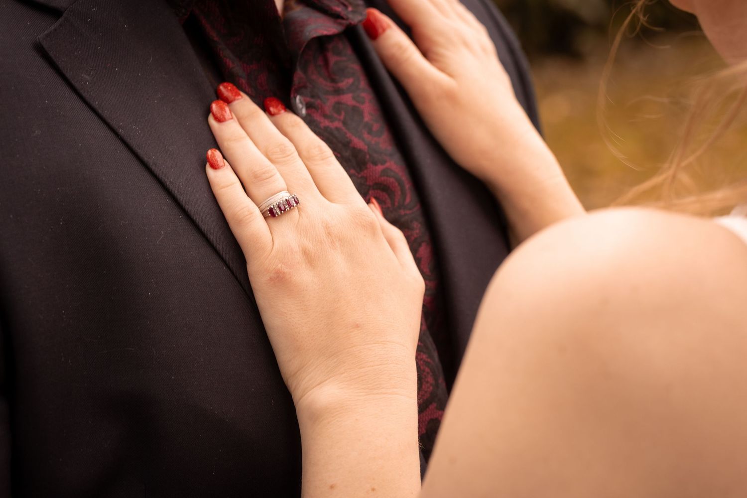 Close-up of a bride's hand with a ring and red nails resting on a groom's dark suit jacket.