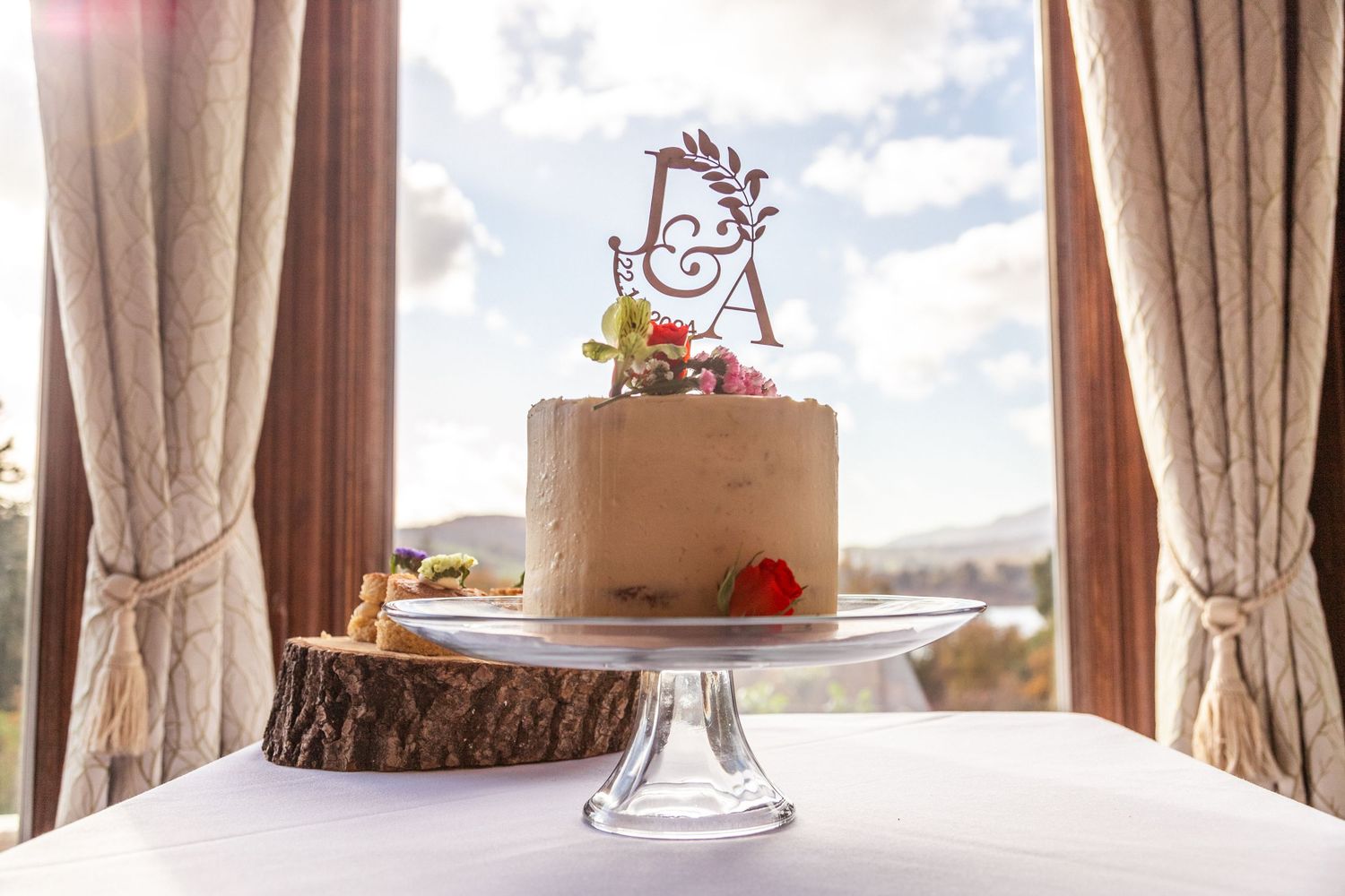 Single-tier wedding cake with monogram topper on glass stand, displayed on rustic wood slice with scenic window backdrop.
