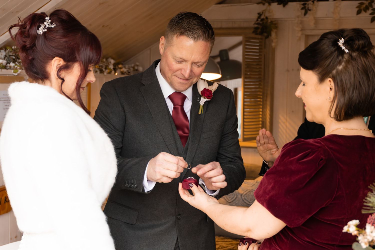 Groom placing wedding ring on brides finger during indoor ceremony, with bridesmaid holding ring box in burgundy dress.