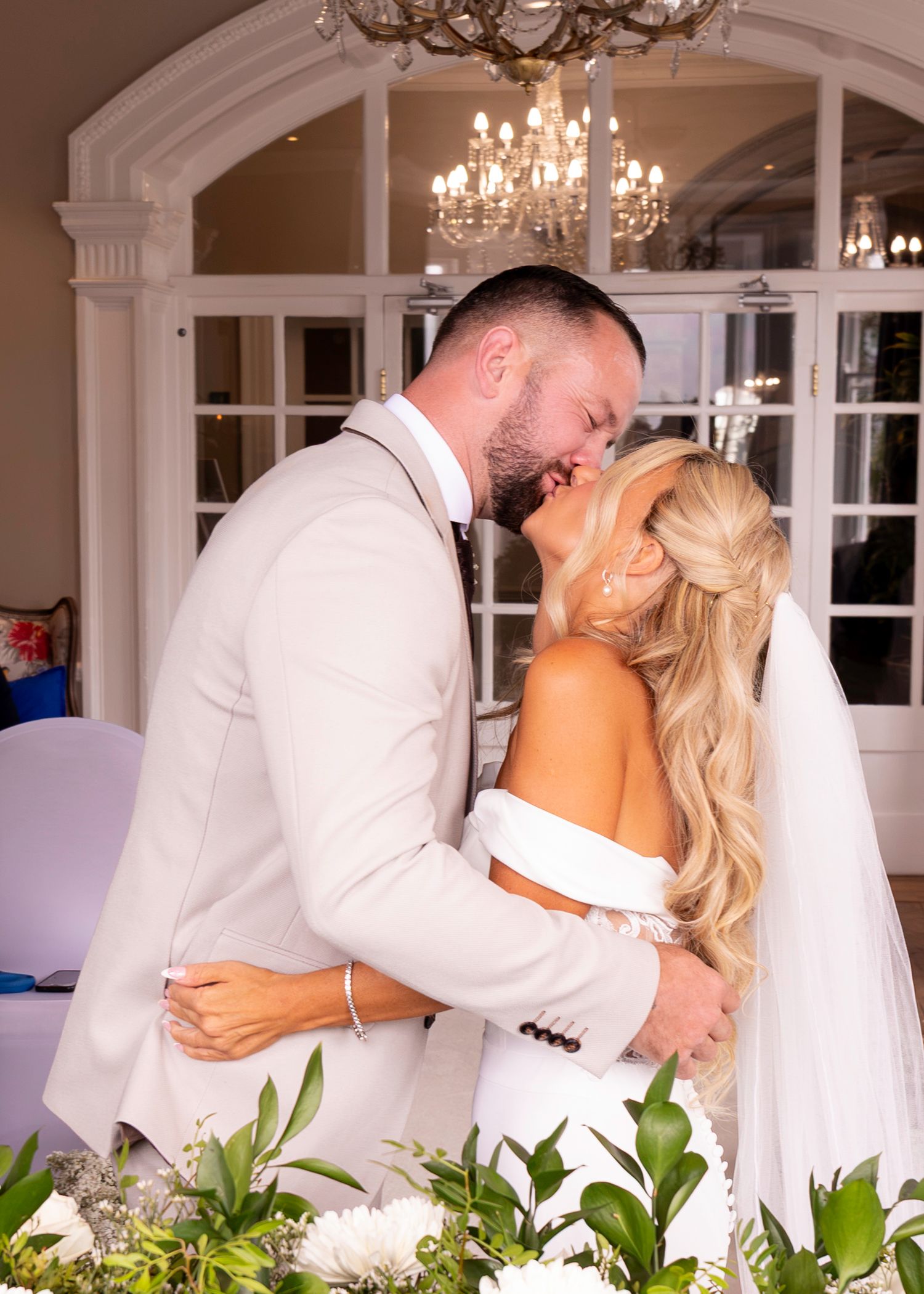 A bride and groom share their first kiss at an elegant wedding venue with a chandelier and French doors.