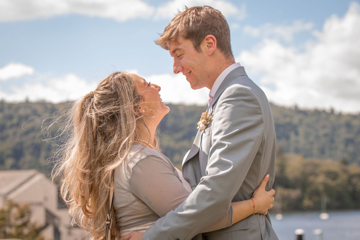 Joyful newlywed couple embracing with lake and hills in background on a bright sunny wedding day.