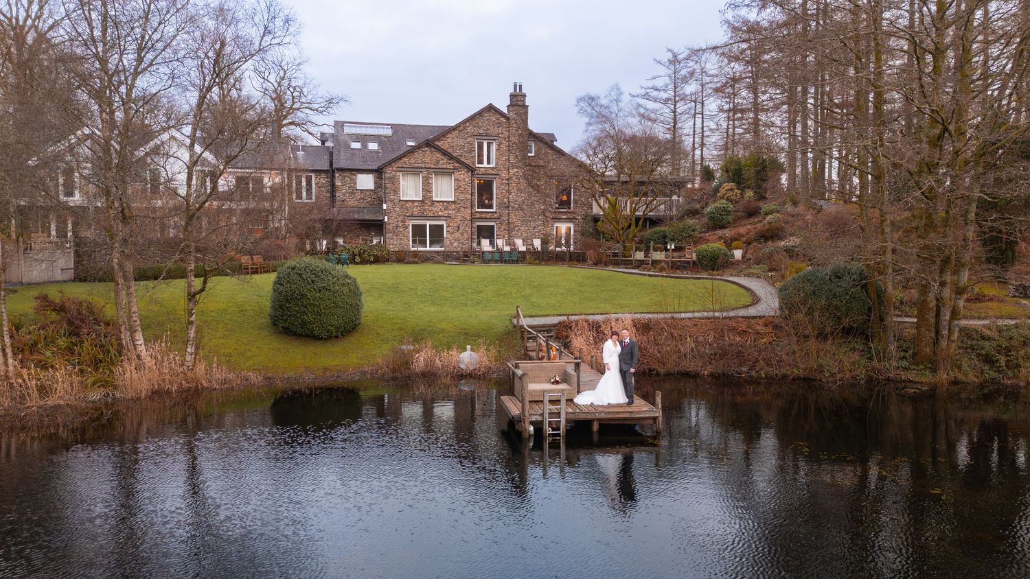 Aerial view of bride and groom standing on a lakeside dock beside a large stone country house.