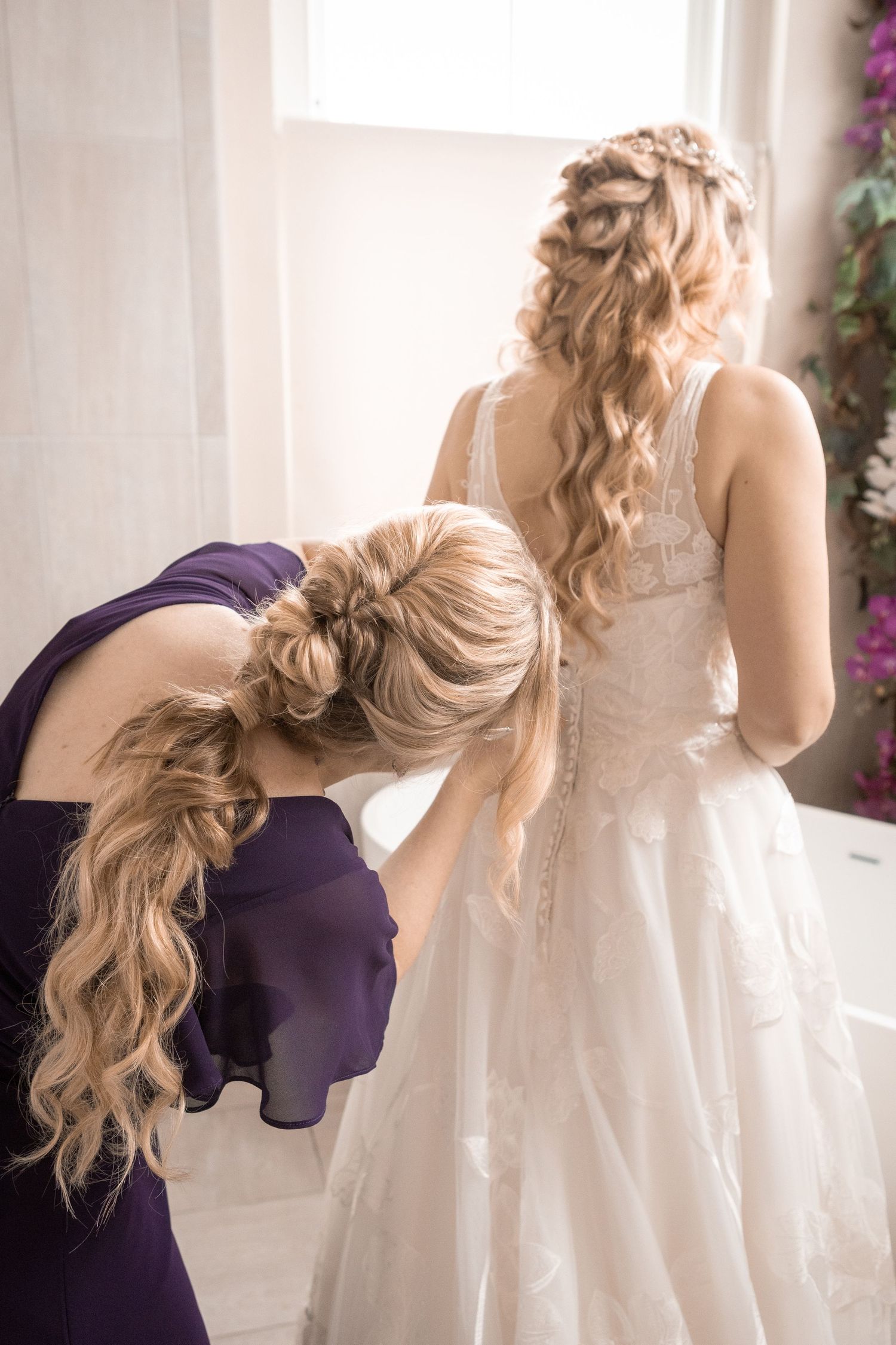 Bridesmaid in purple dress fastens the back of a bride's lace wedding gown near a bright window.