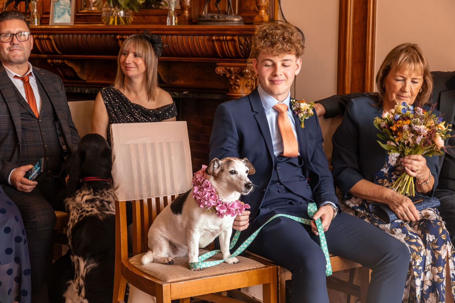 Dog sitting on chair wearing floral collar at wedding ceremony with guests.