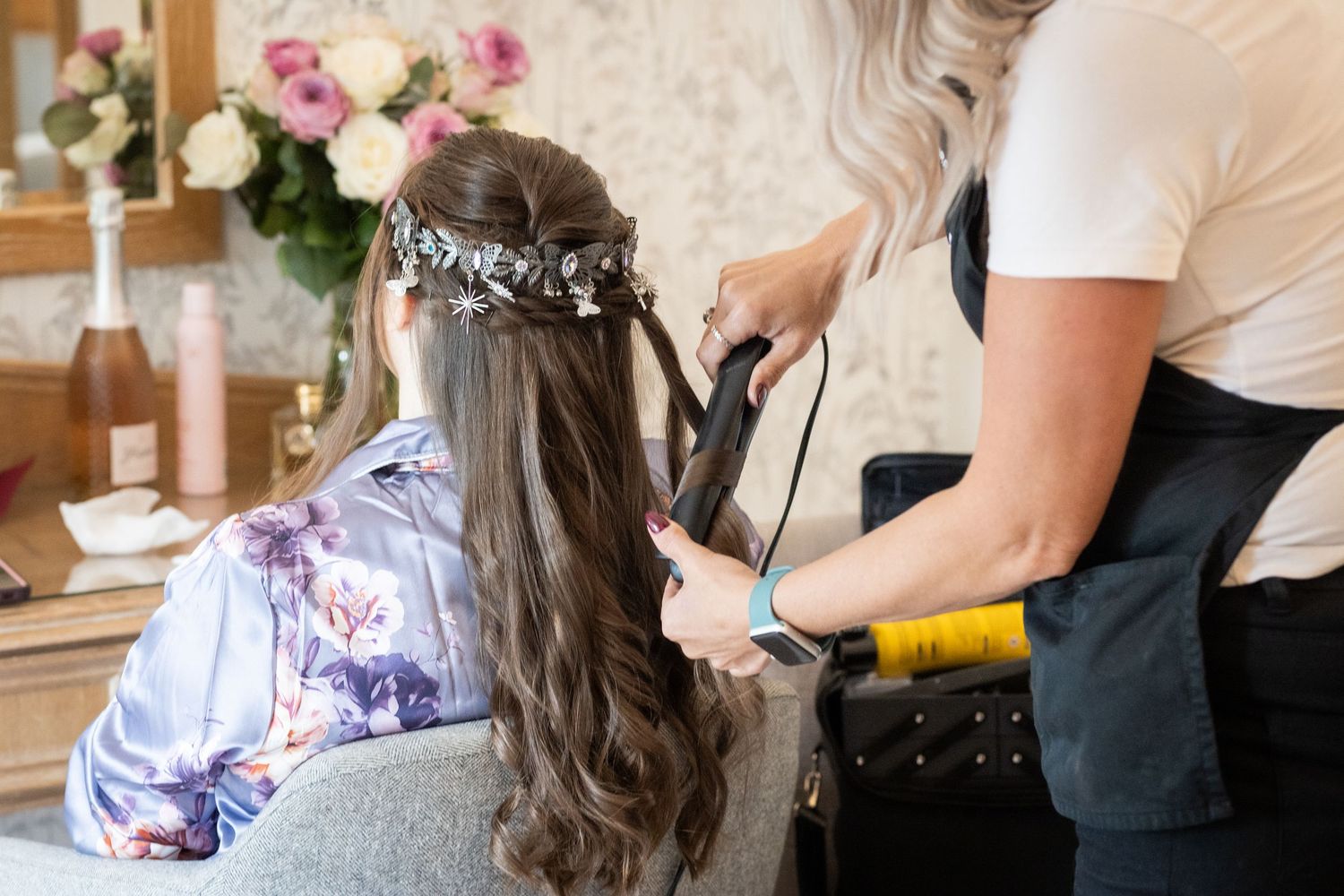 Hairstylist curling long brown hair of a bride wearing a floral crown and silk robe for wedding preparation.