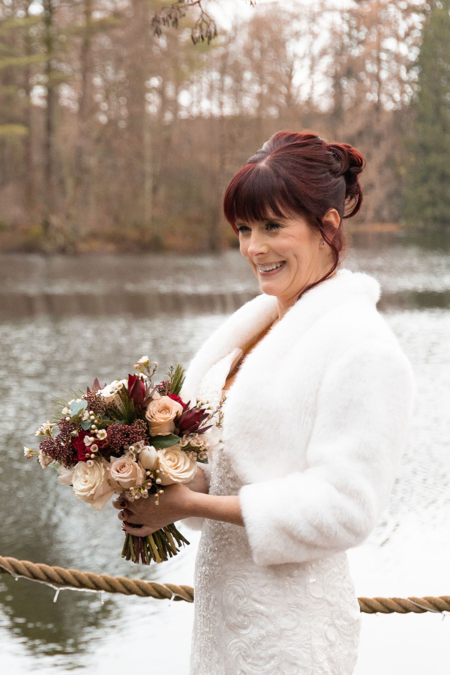 Smiling bride in white fur coat holding bouquet of roses by a lake in winter woodland setting.