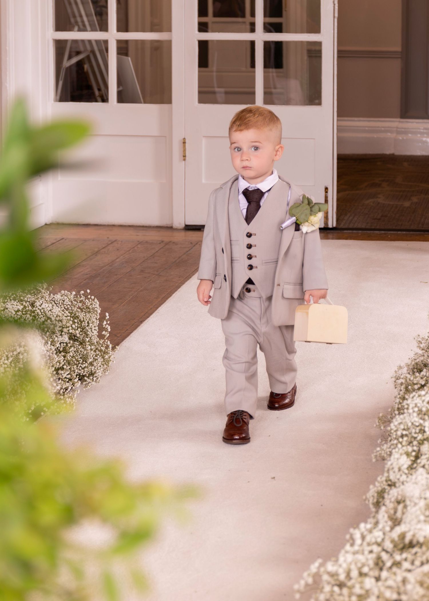Young ring bearer in grey suit carries box down wedding aisle.