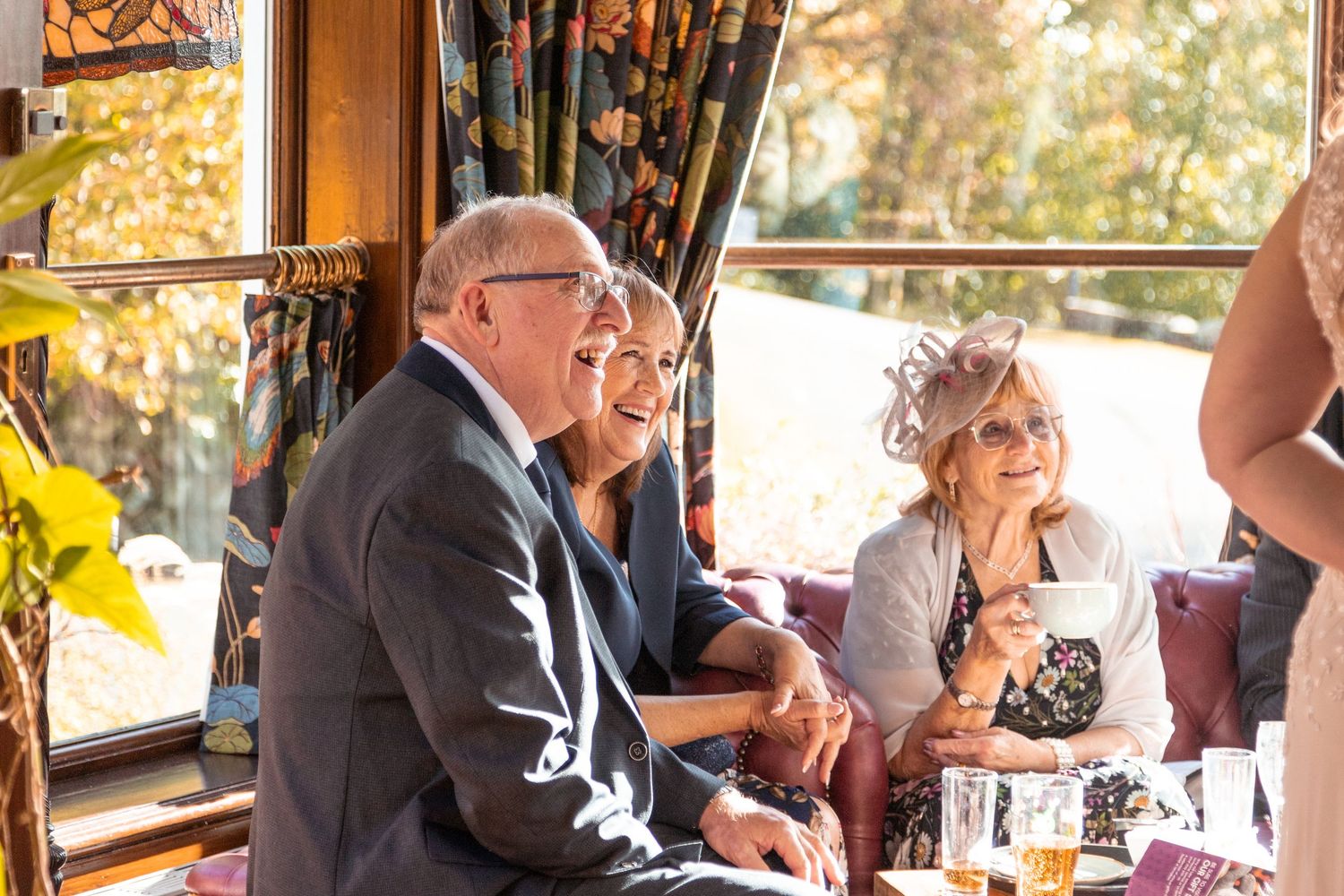 Wedding guests laughing and socializing indoors with autumn views through decorative windows.