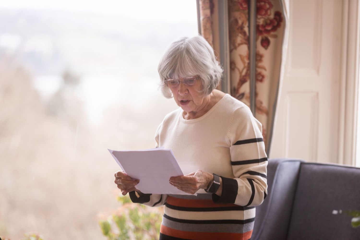Senior woman with gray hair and glasses reading a document by a bright window in a cozy home.