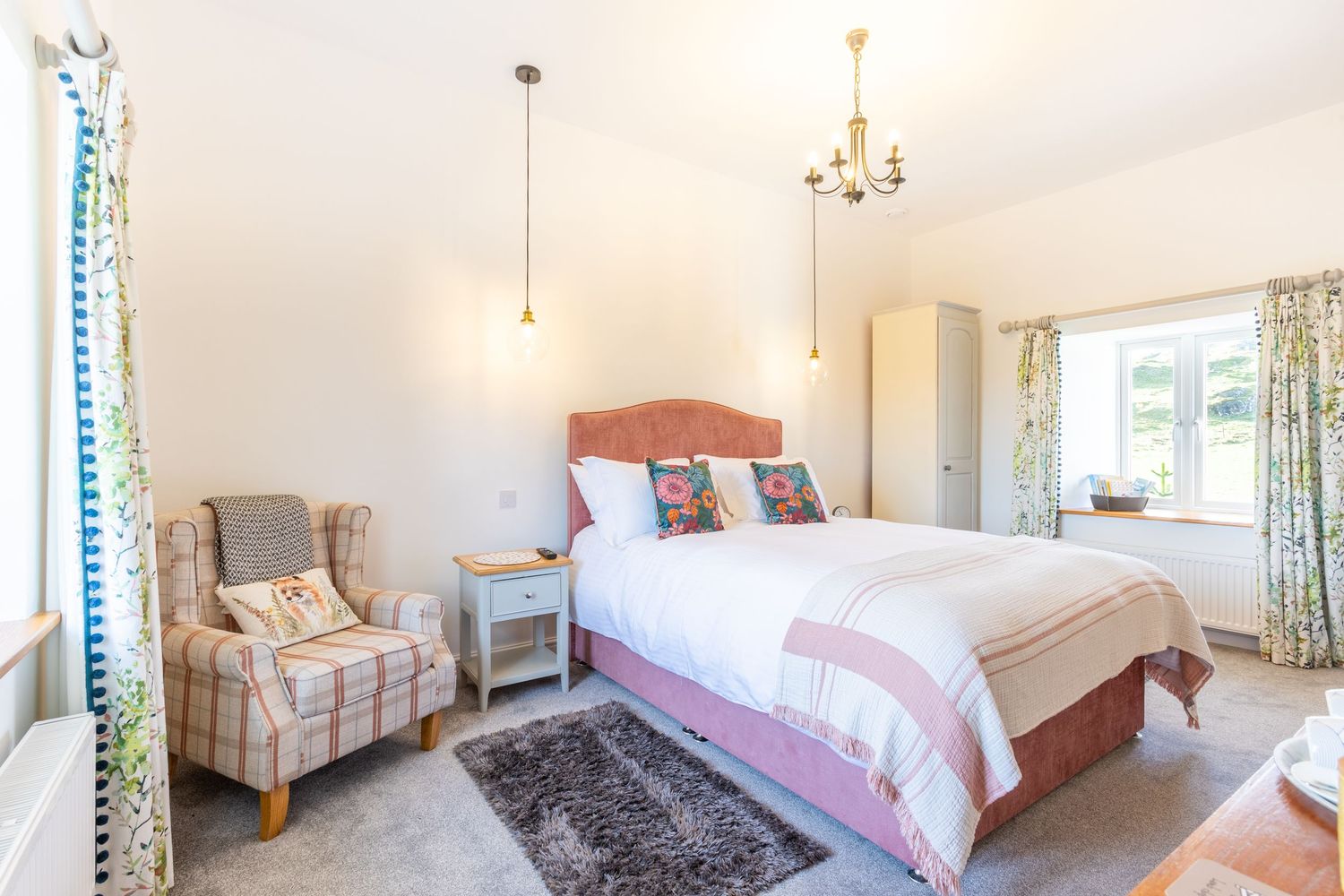 Spacious country bedroom with pink headboard, tartan armchair, chandelier, and natural light from large window.