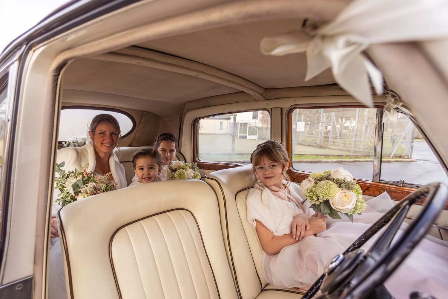 Bride and flower girls seated inside a vintage cream-colored classic wedding car interior.