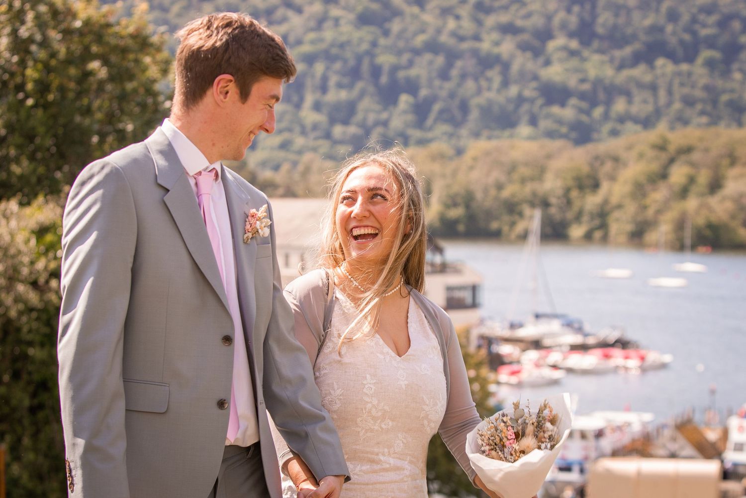 A laughing bride holding a bouquet stands with her groom overlooking a scenic harbor on their wedding day.