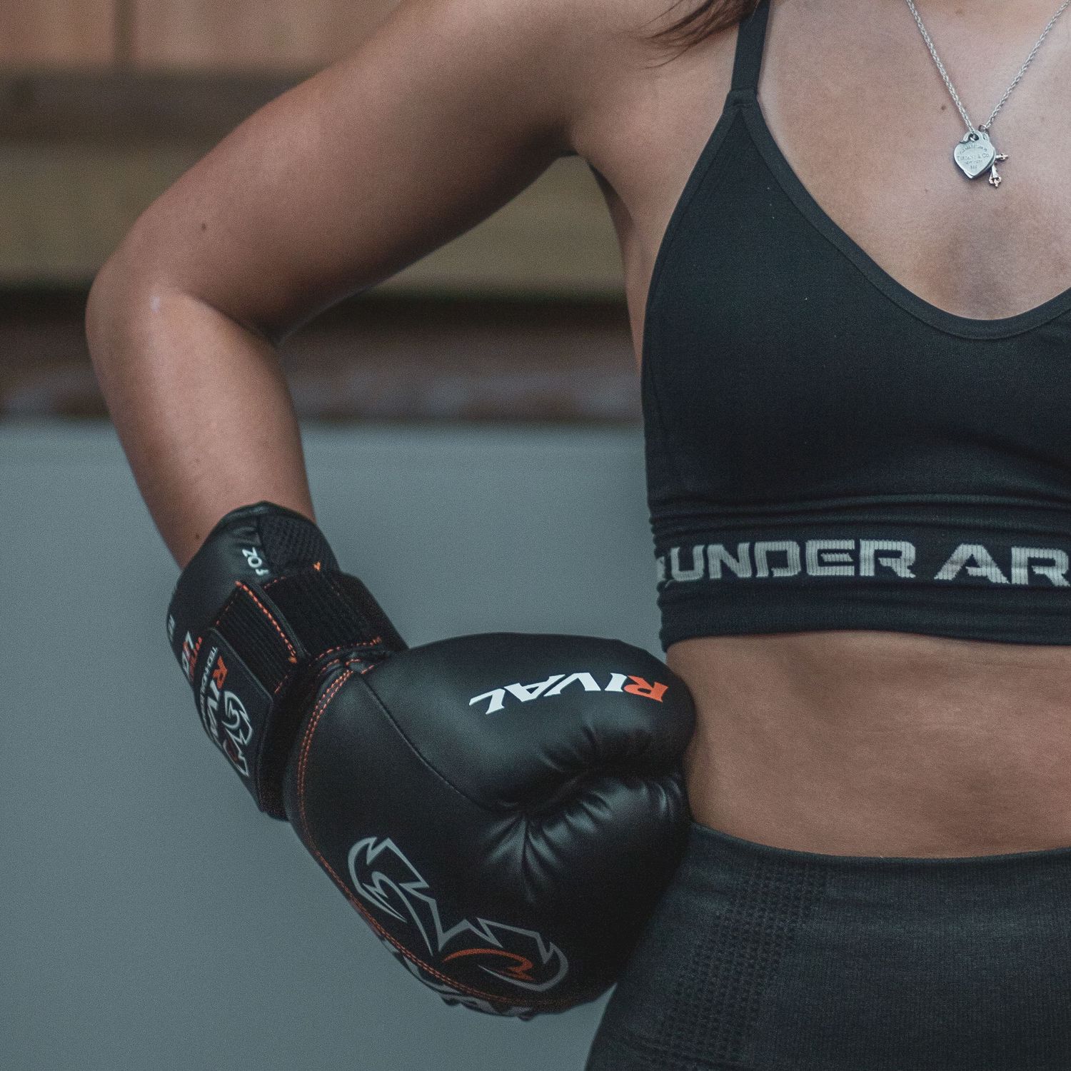 Female boxer in Under Armour sports bra poses with black Rival boxing gloves in a gym.