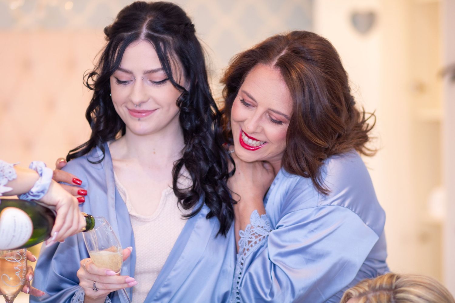 Two women in blue satin robes smile while pouring champagne during bridal preparations.