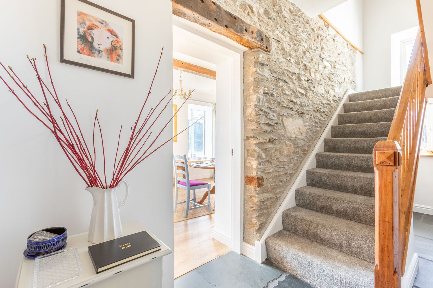 Bright hallway with exposed stone wall, carpeted staircase, wooden banister, and decorative vase with red stems.