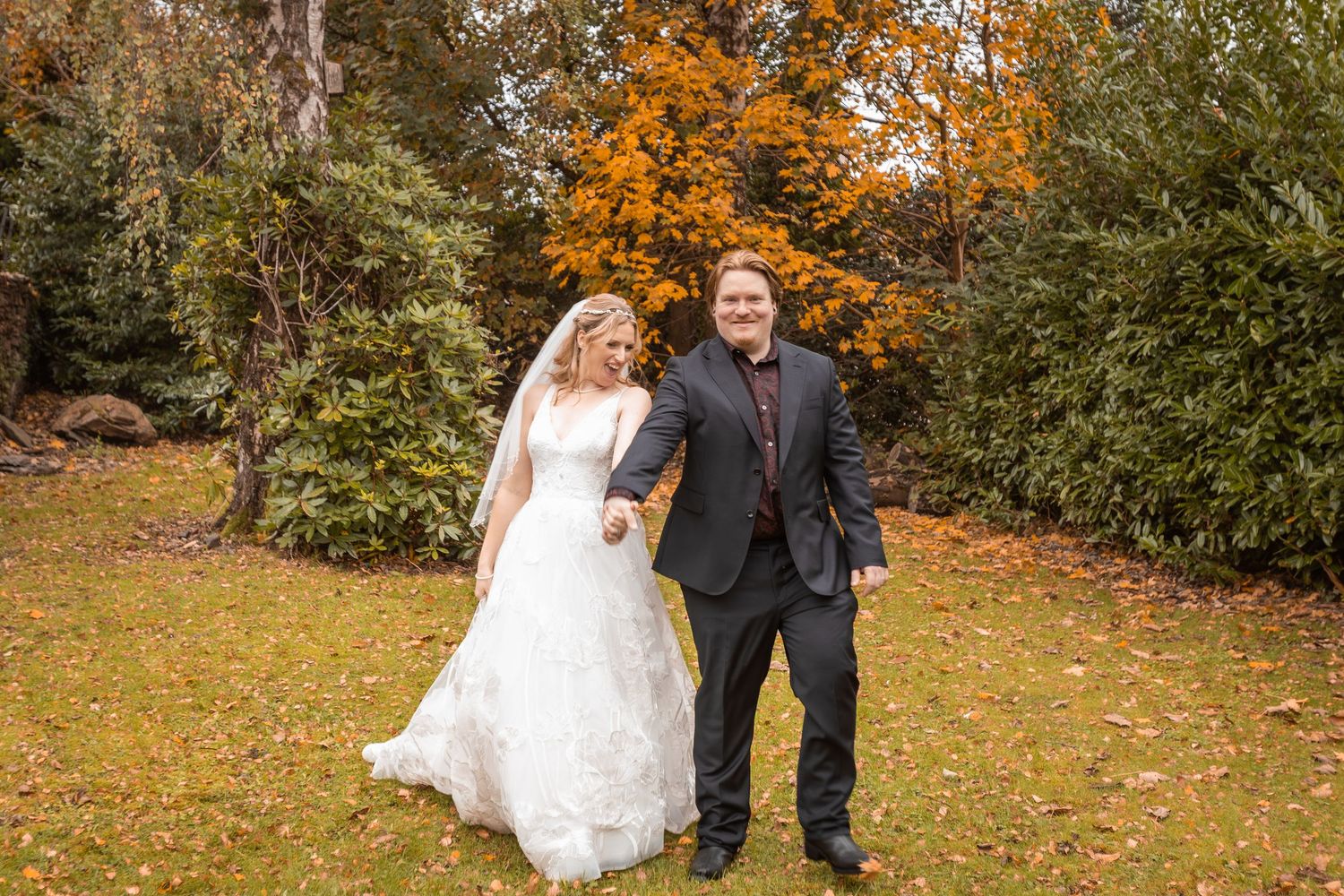 Bride and groom walking hand in hand on autumn lawn surrounded by colorful fall foliage during outdoor wedding photo session.