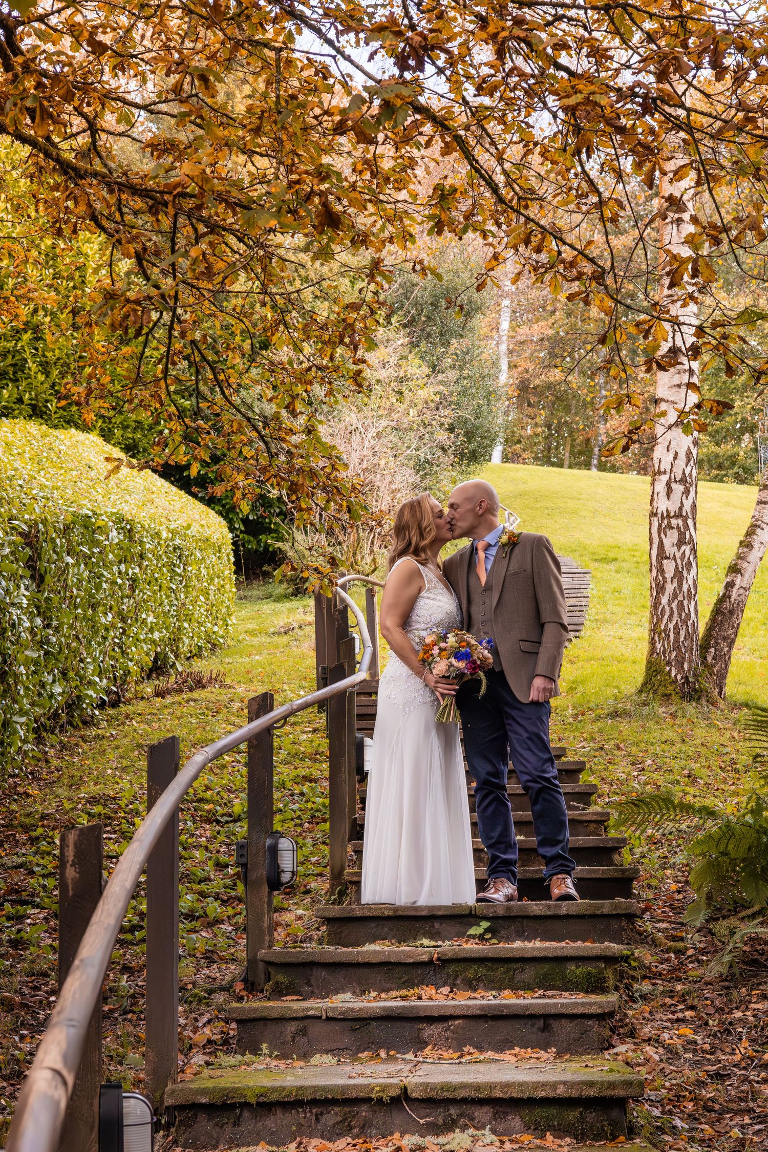 Bride and groom kissing on wooden steps surrounded by golden autumn foliage in a lush garden setting.
