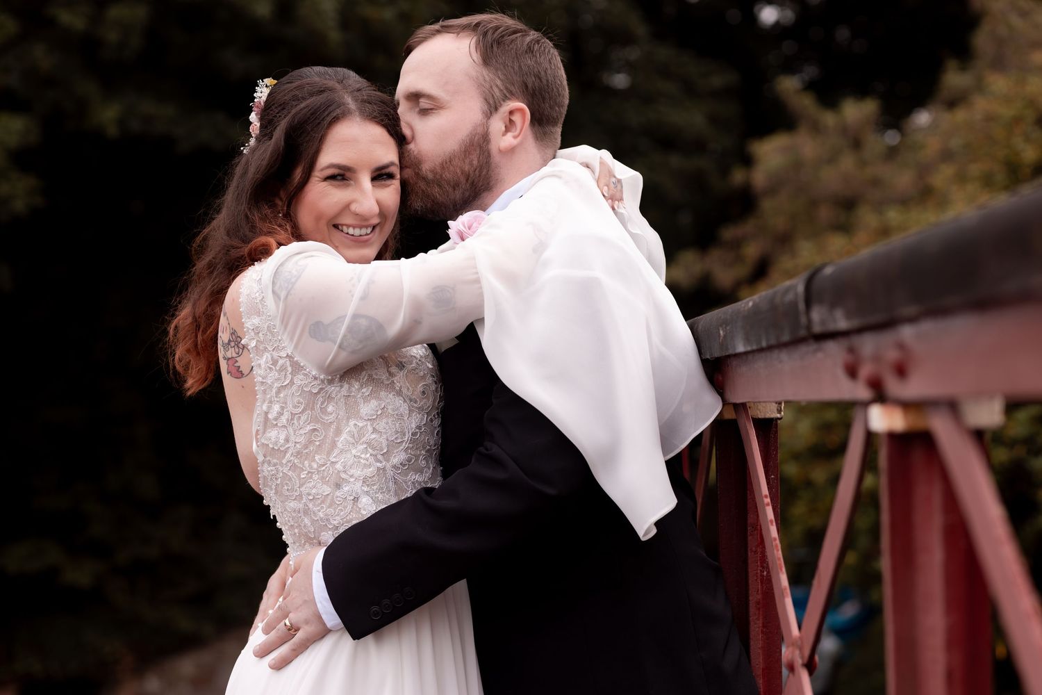 A bride in lace gown embraces groom in white tuxedo jacket on a bridge, smiling joyfully at their wedding.