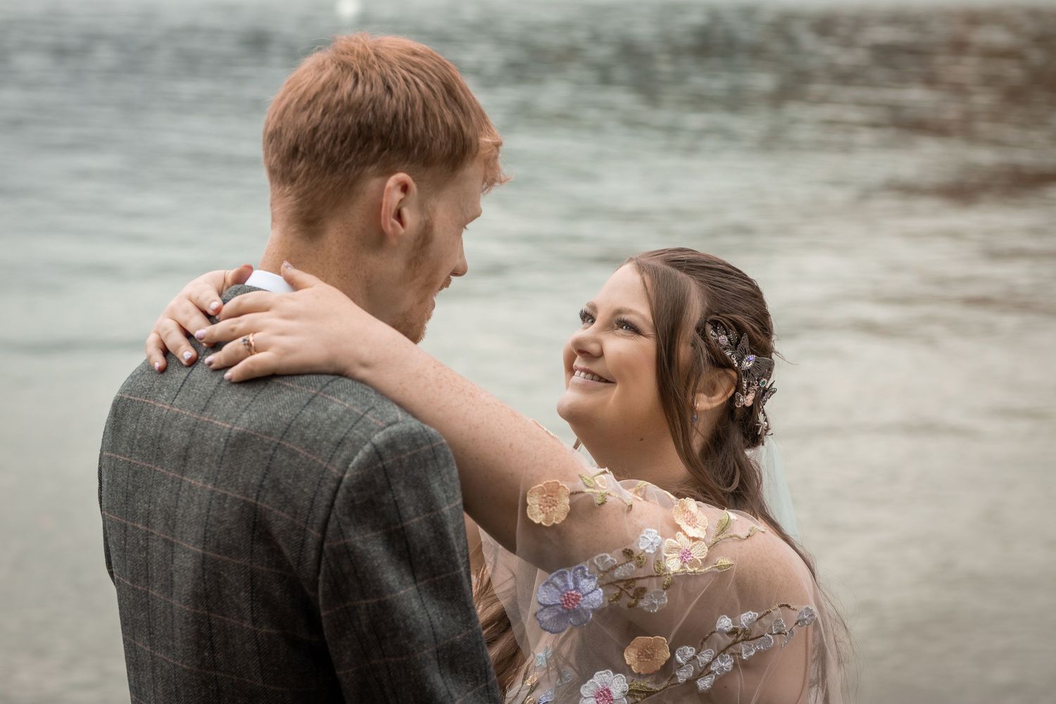 Bride in floral gown smiles at groom in plaid suit near waterfront in romantic outdoor wedding portrait.