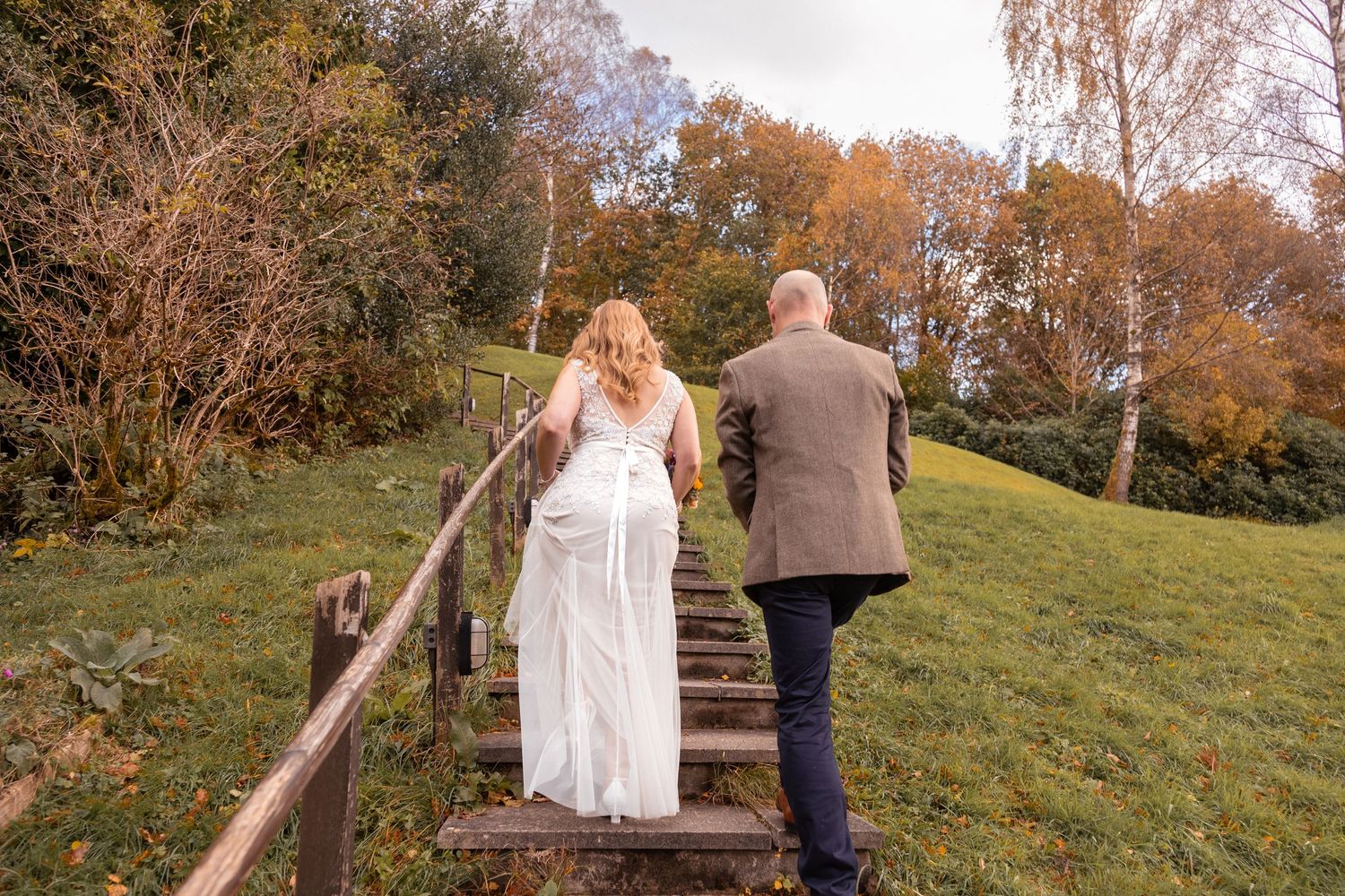 A bride in a white gown and groom in tweed jacket walk up wooden steps in autumn.