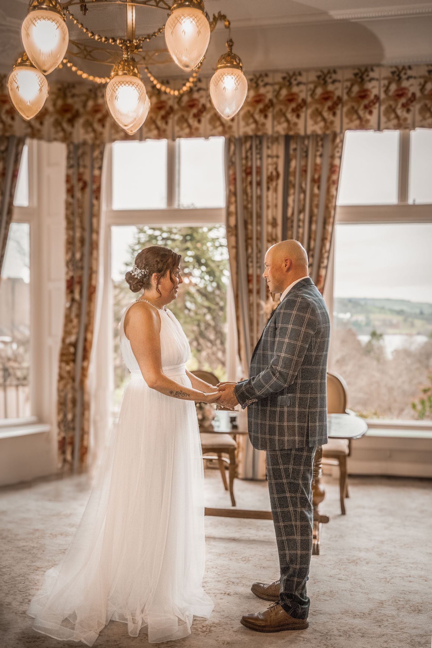 Bride and groom holding hands during indoor wedding ceremony in elegant room with chandelier and floral curtains.
