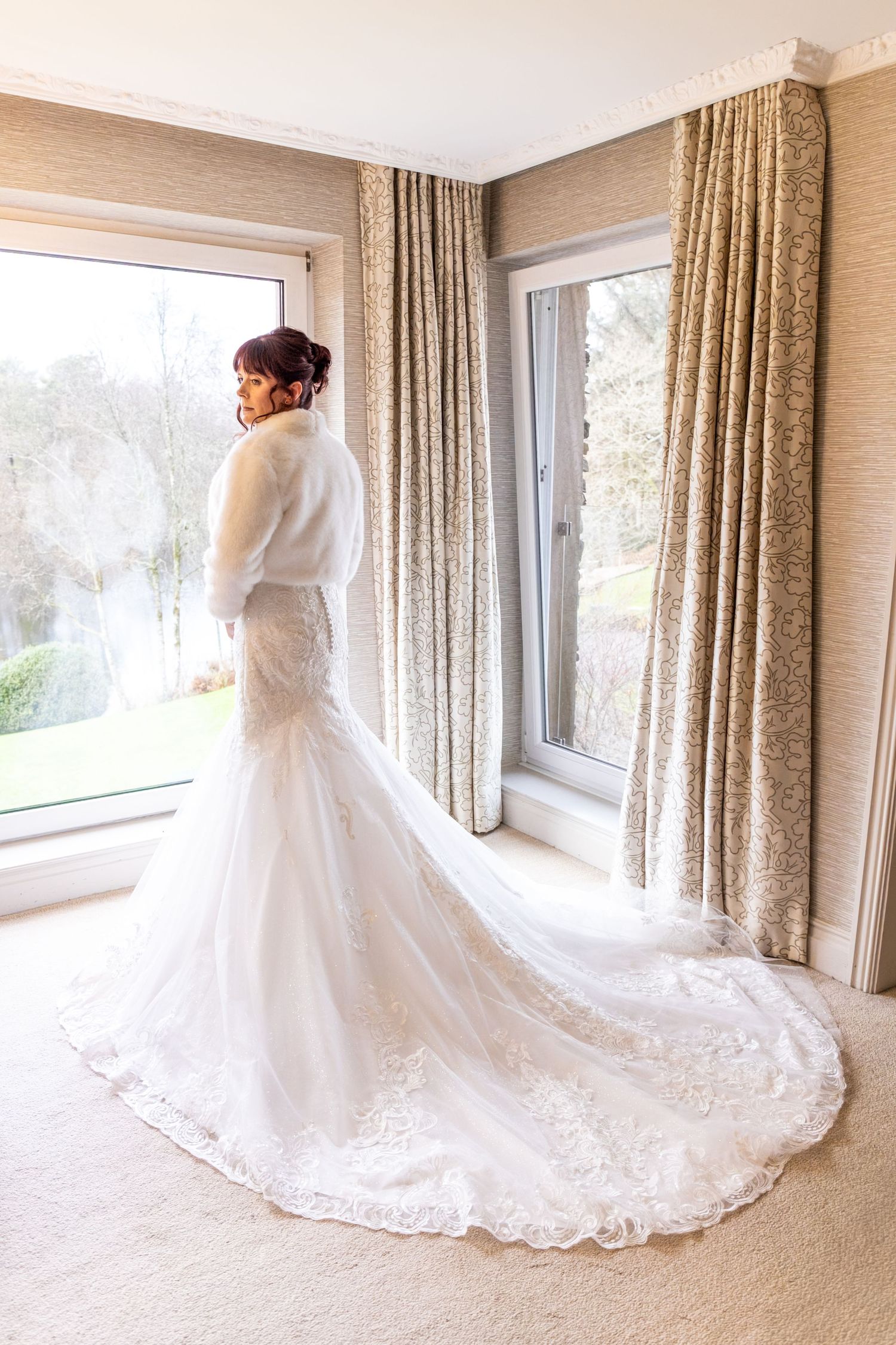 Bride in white lace mermaid wedding gown with fur shawl and long train standing by window in elegant room.