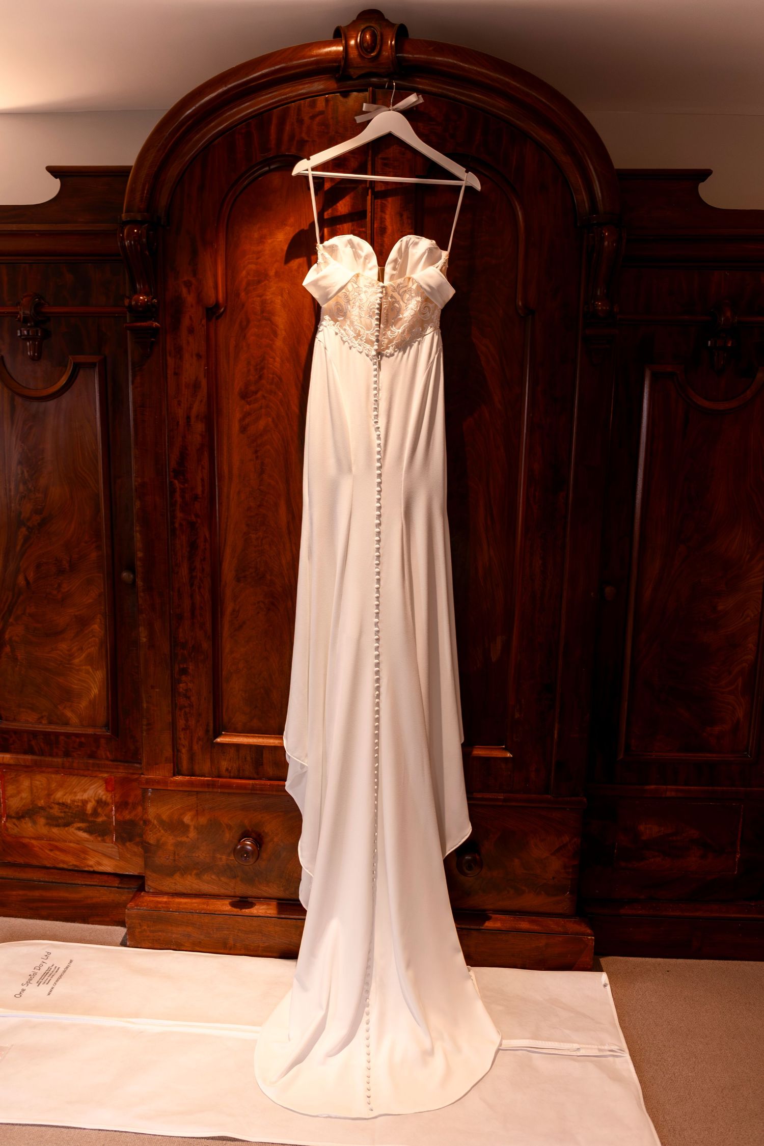 Elegant strapless white wedding gown with beaded bodice and button back, displayed on hanger against ornate wooden armoire.