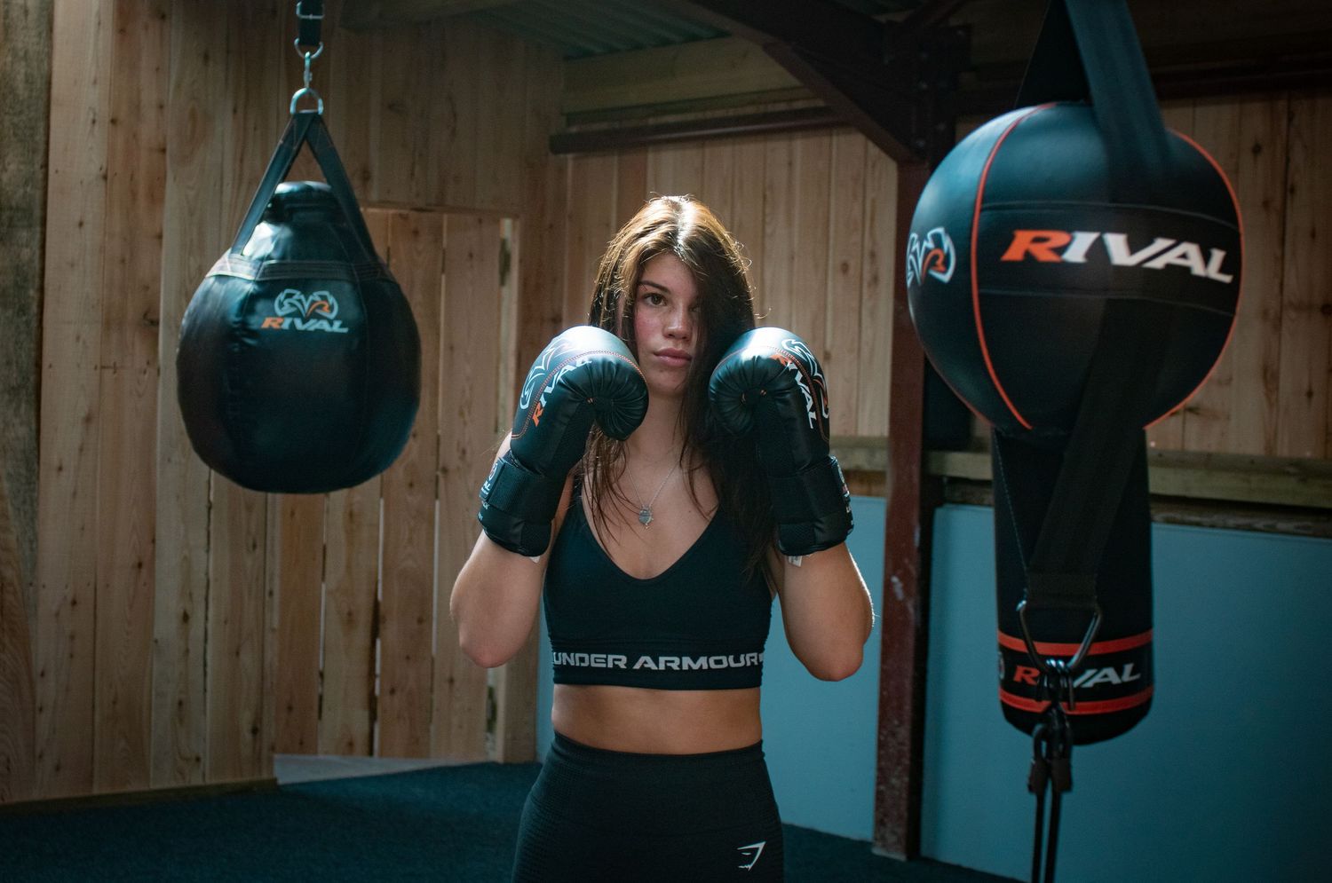 Female boxer in black gloves training between two Rival punching bags in a rustic boxing gym.