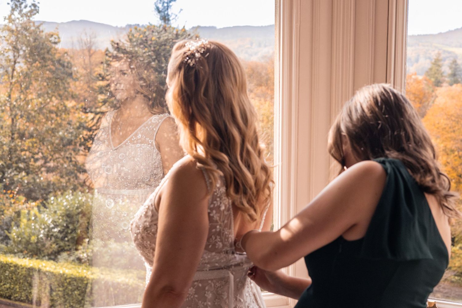 Bridesmaid helping bride with dress by window with autumn foliage views.