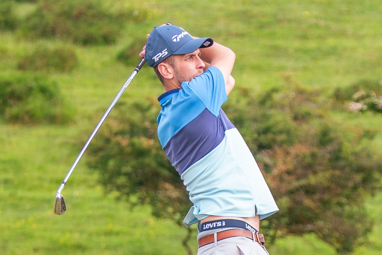Golfer in blue shirt completing a powerful swing on a lush green golf course.