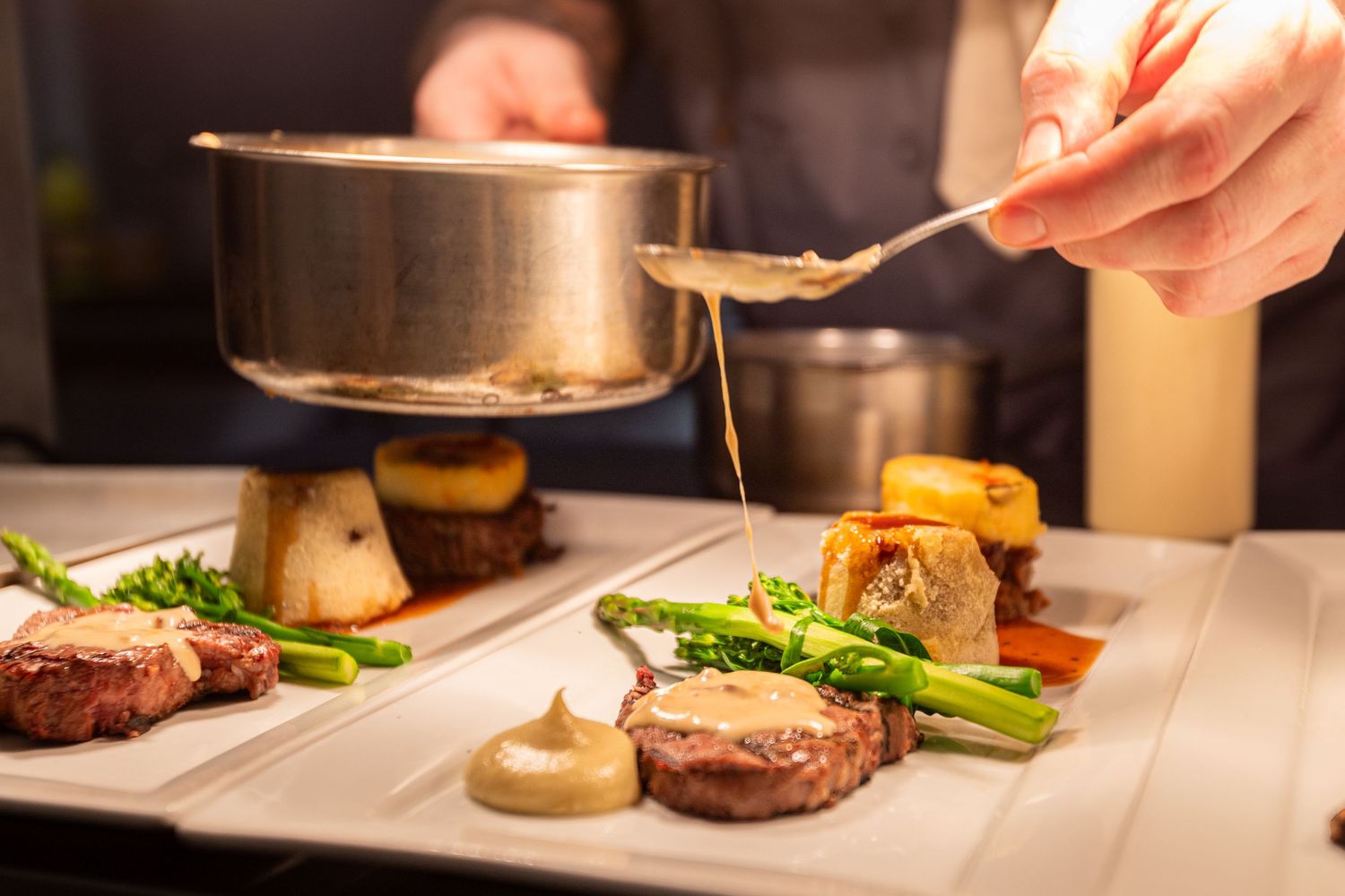 Chef pouring sauce over an elegant plated dish with steak and asparagus.