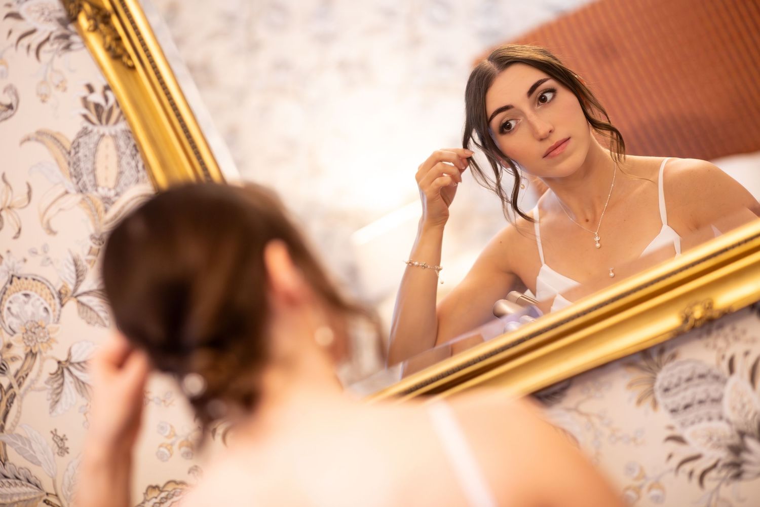 Young woman in white dress adjusting earring while looking at her reflection in an ornate gold-framed mirror.