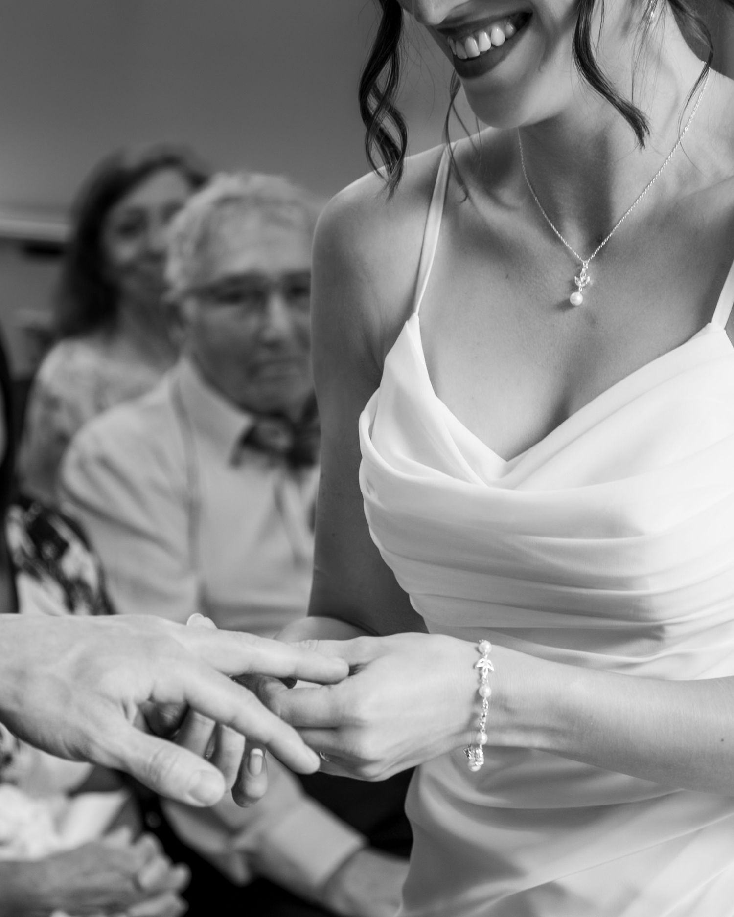 Black and white photo of bride smiling during ring exchange ceremony, wearing elegant white gown and pearl bracelet.