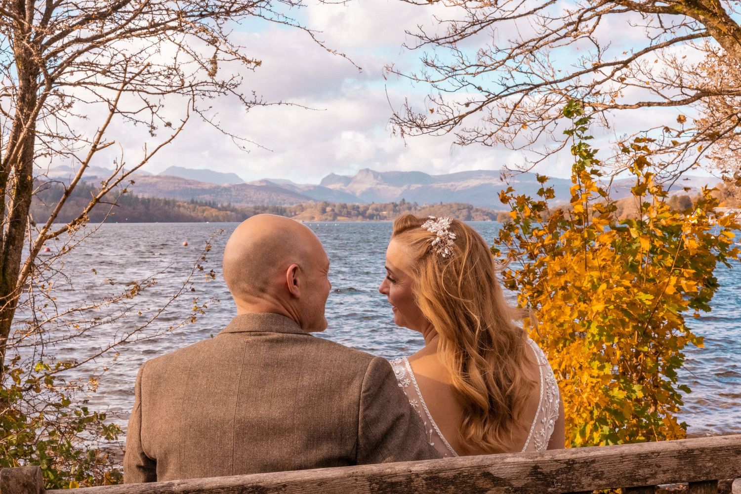 Bride and groom sitting on a bench overlooking a scenic lake surrounded by autumn trees.