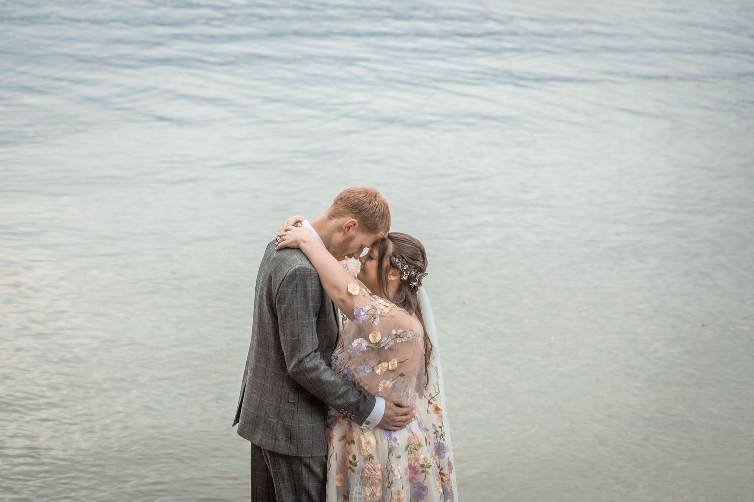 Couple embracing romantically by the water's edge, bride in floral dress with flowers in hair, groom in gray suit.
