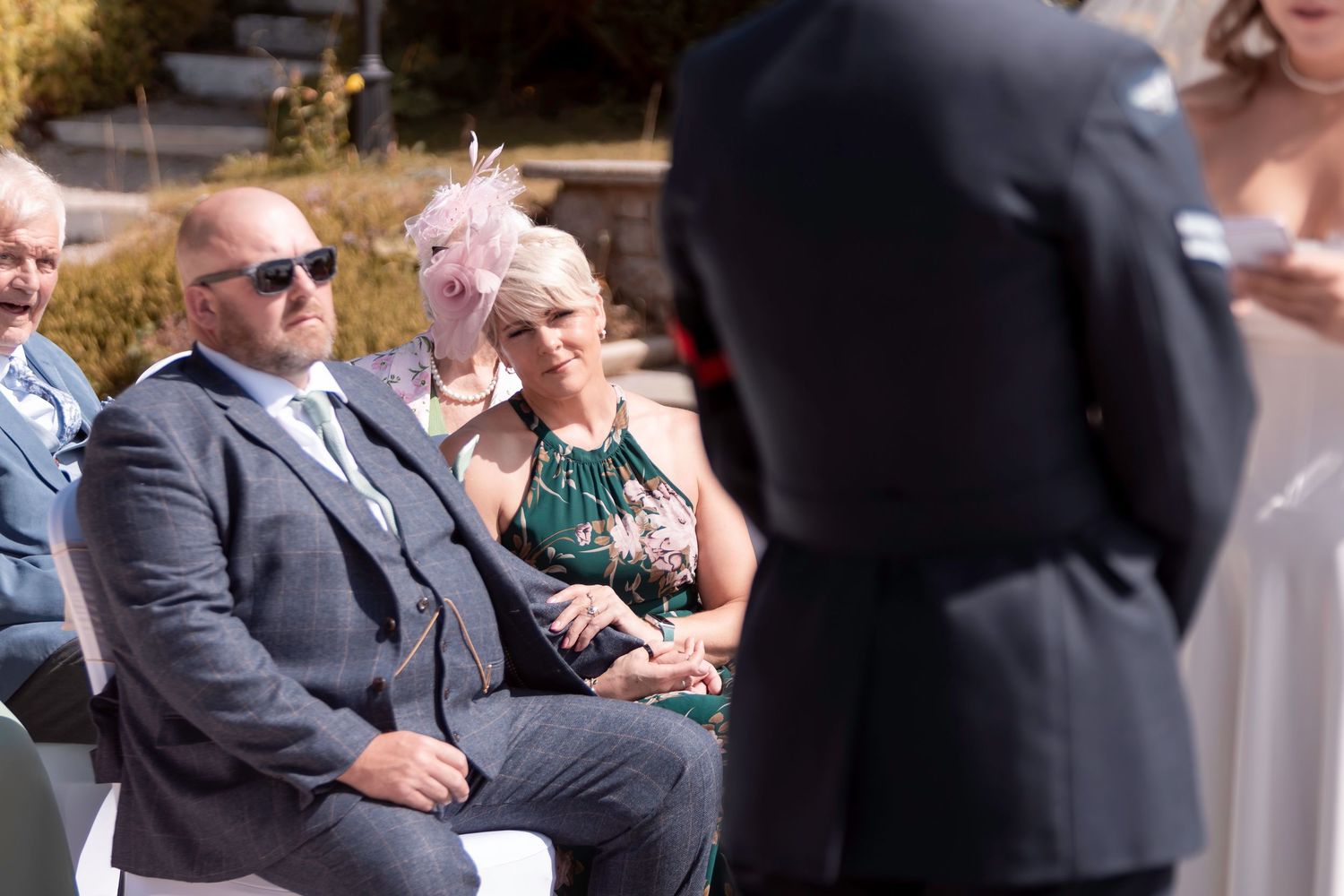 Wedding guests seated outdoors, smiling during a ceremony, with a woman in a pink fascinator and man in a grey suit.
