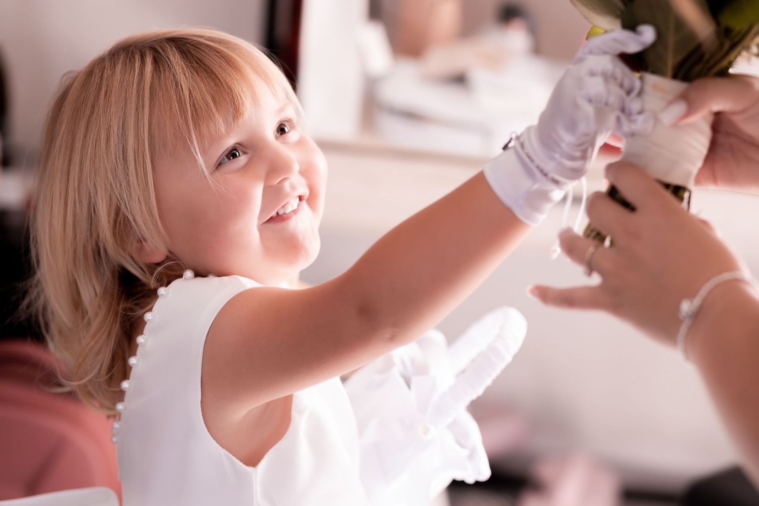 A smiling young girl in a white dress and gloves reaches out to receive a floral arrangement at a wedding ceremony.