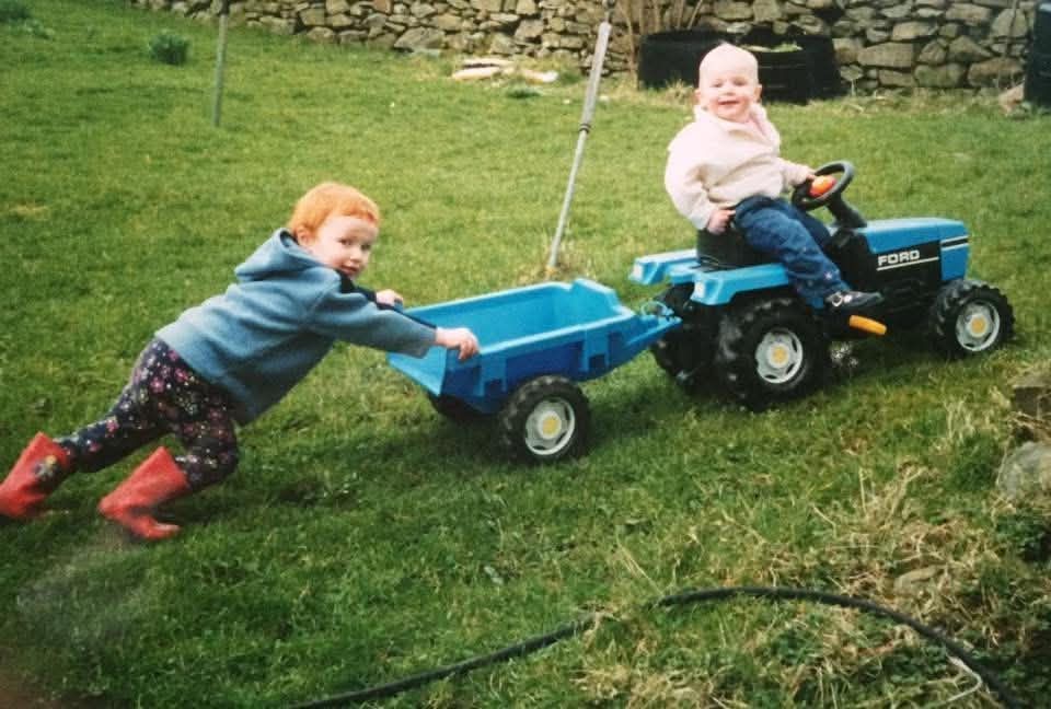 Two young children playing outdoors with a blue pedal tractor and trailer toy on a green lawn.