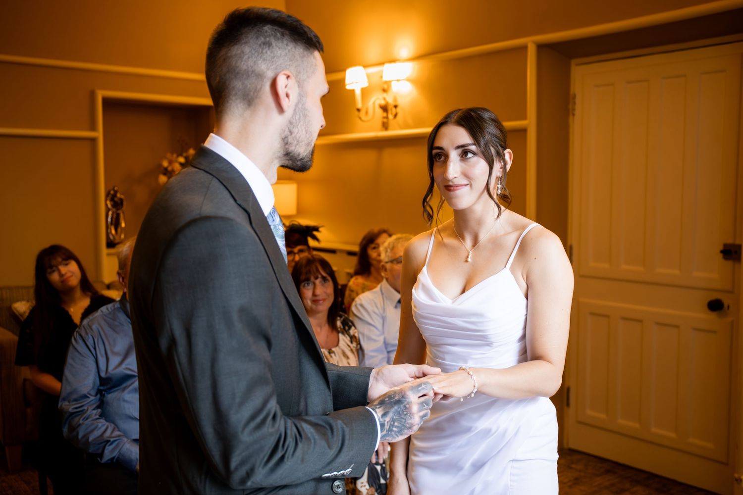 Bride and groom exchange rings during intimate wedding ceremony, guests seated in warmly lit venue background.
