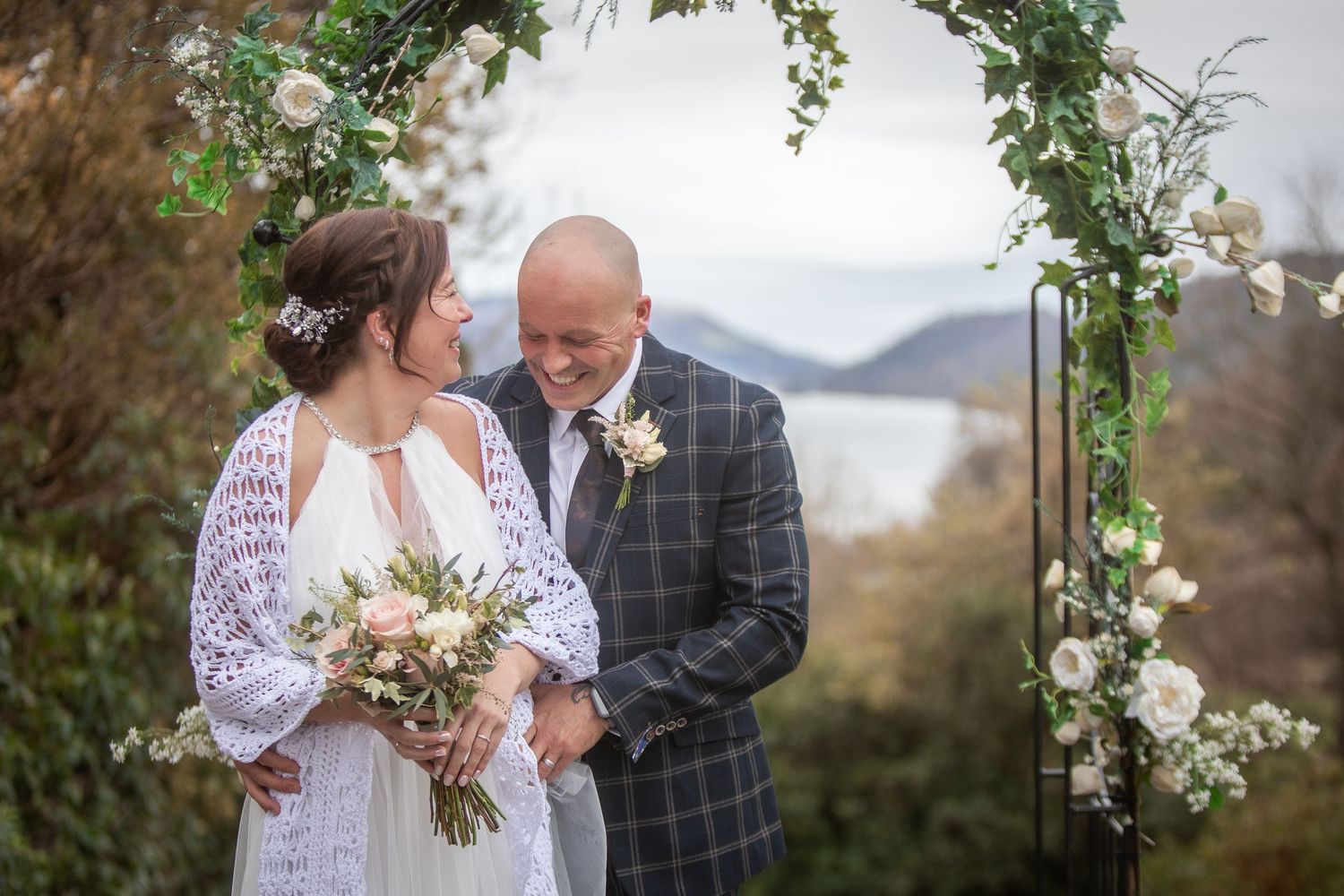 Bride and groom share a tender moment under a floral arch with a stunning lake and mountain backdrop.