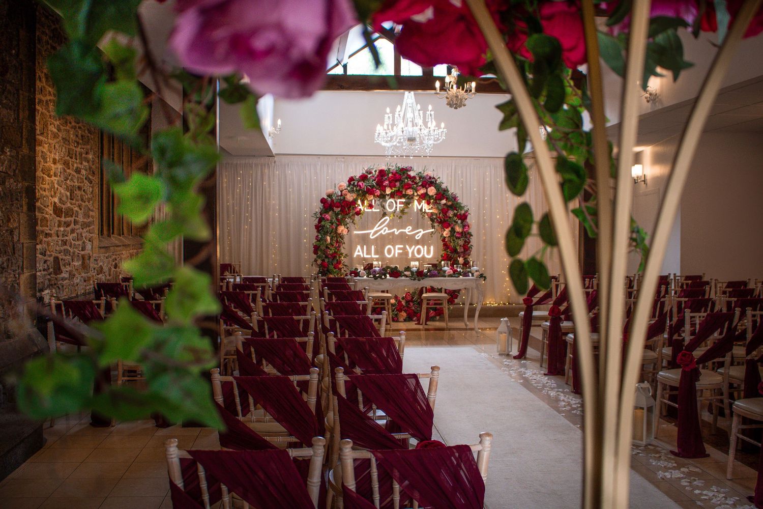 Romantic wedding venue with burgundy chairs, floral arch, and glowing neon sign reading All of Me Loves All of You.