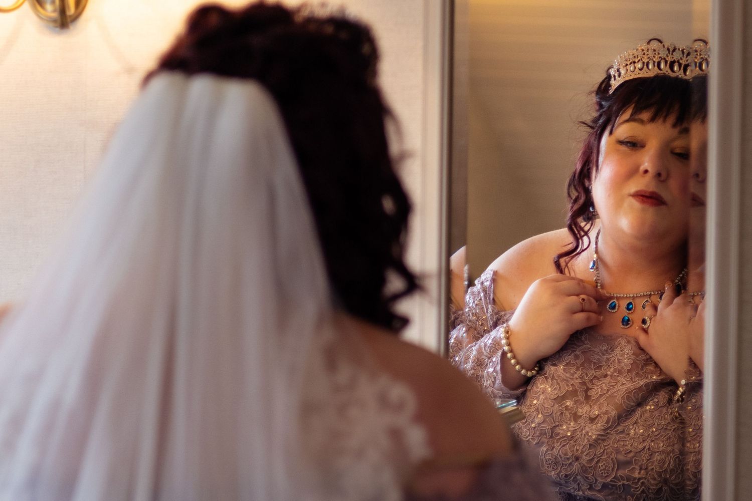A bride wearing a tiara and lace dress adjusts her necklace while looking in a mirror before her wedding.
