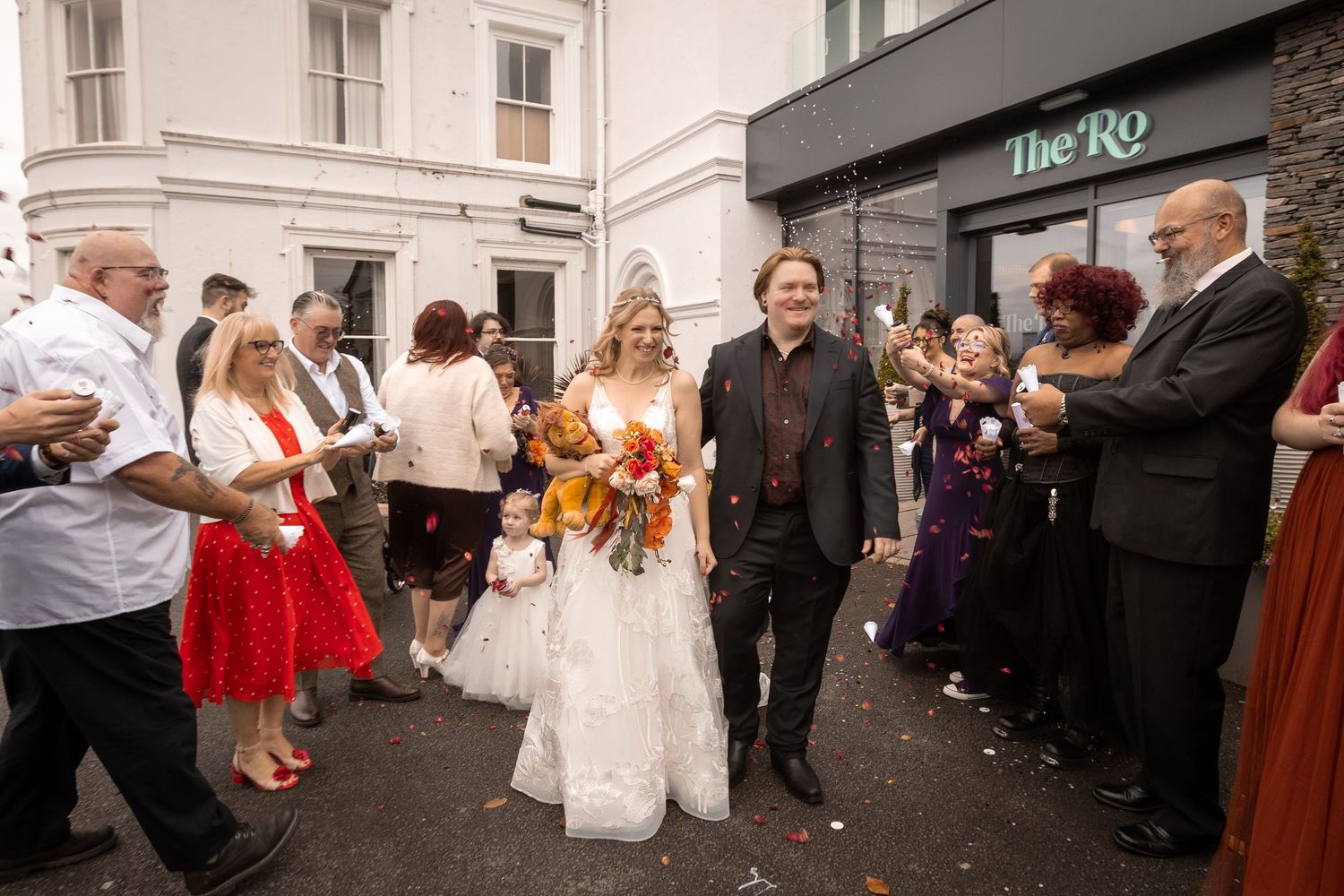 Newlywed couple walks through cheering guests outside The Ro venue, bride in white gown holding orange bouquet.