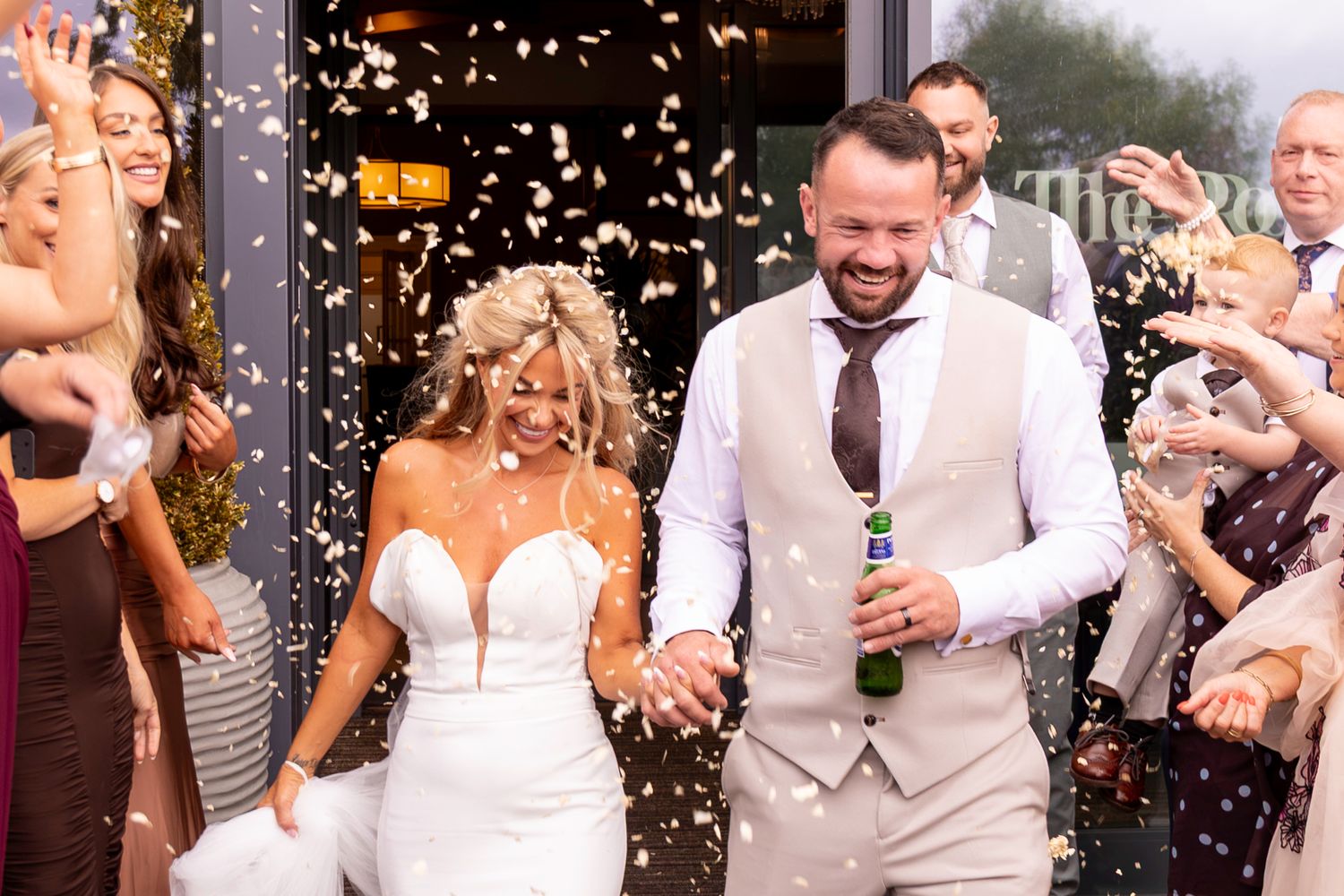 Joyful newlyweds exit wedding venue through confetti shower, bride in white gown, groom in tan suit holding champagne bottle.