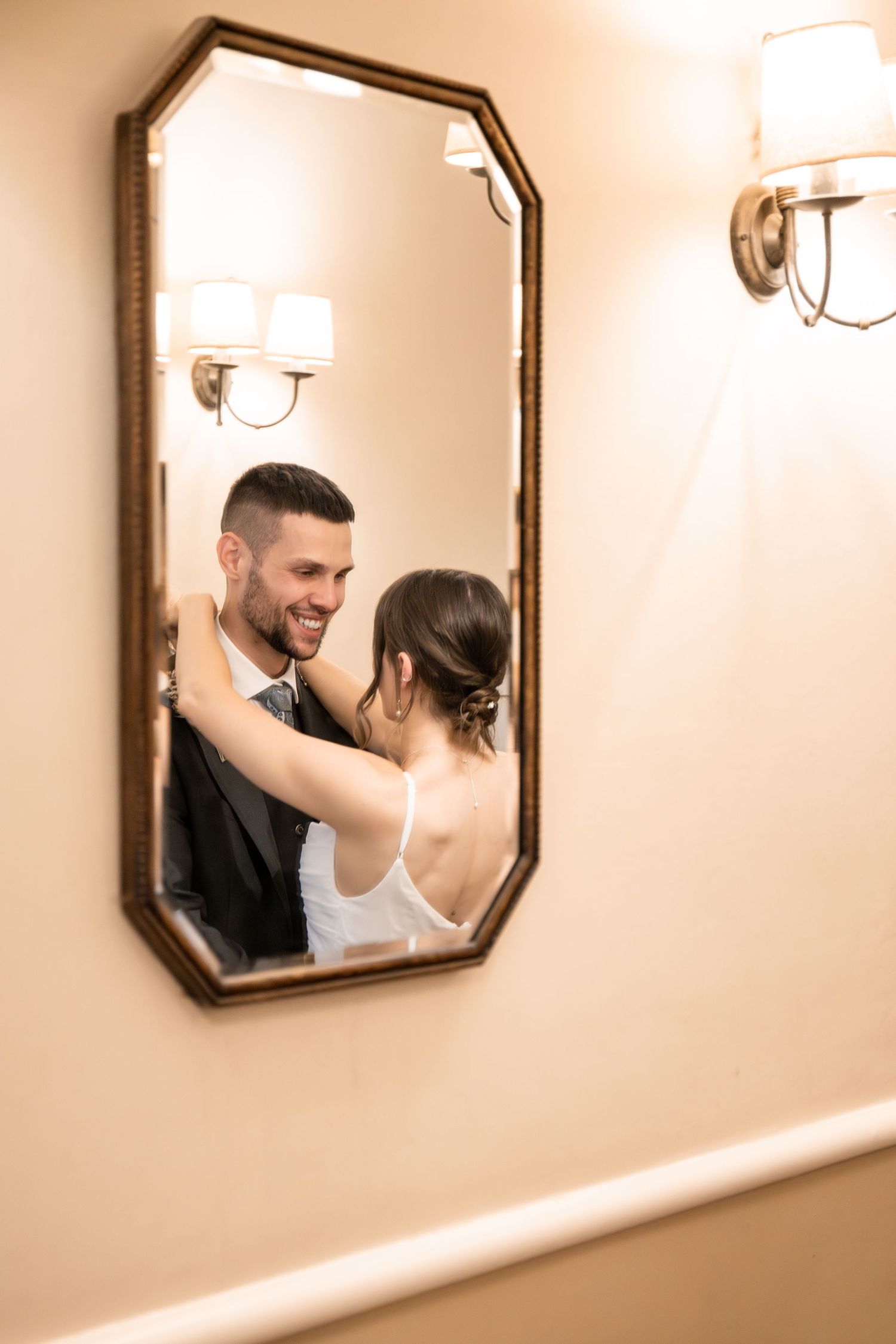 Bride and groom share a romantic embrace reflected in an ornate gold mirror on a beige wall.