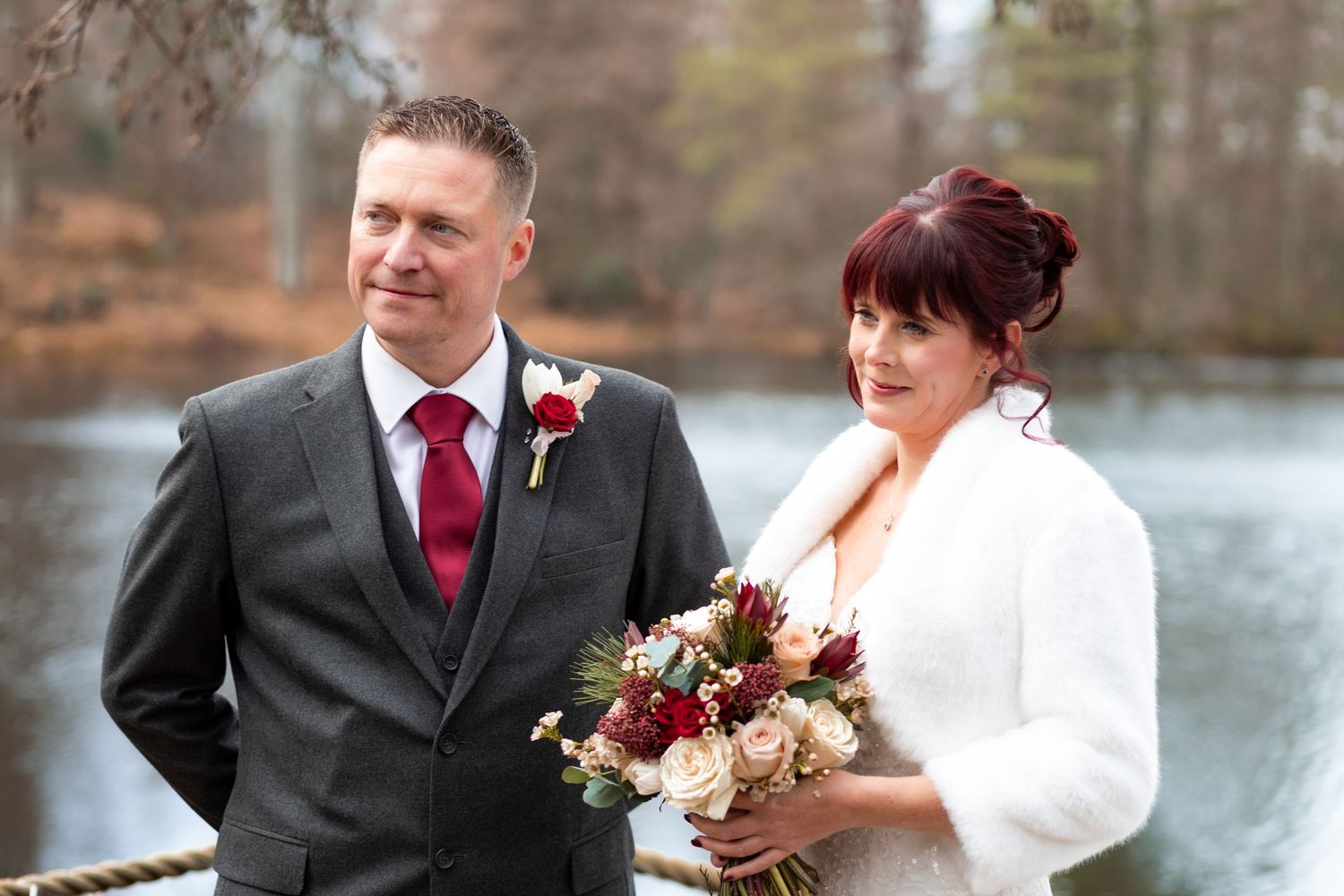 Bride and groom posing outdoors near a lake, bride holding a red and pink bouquet, groom wearing a gray suit with red tie.